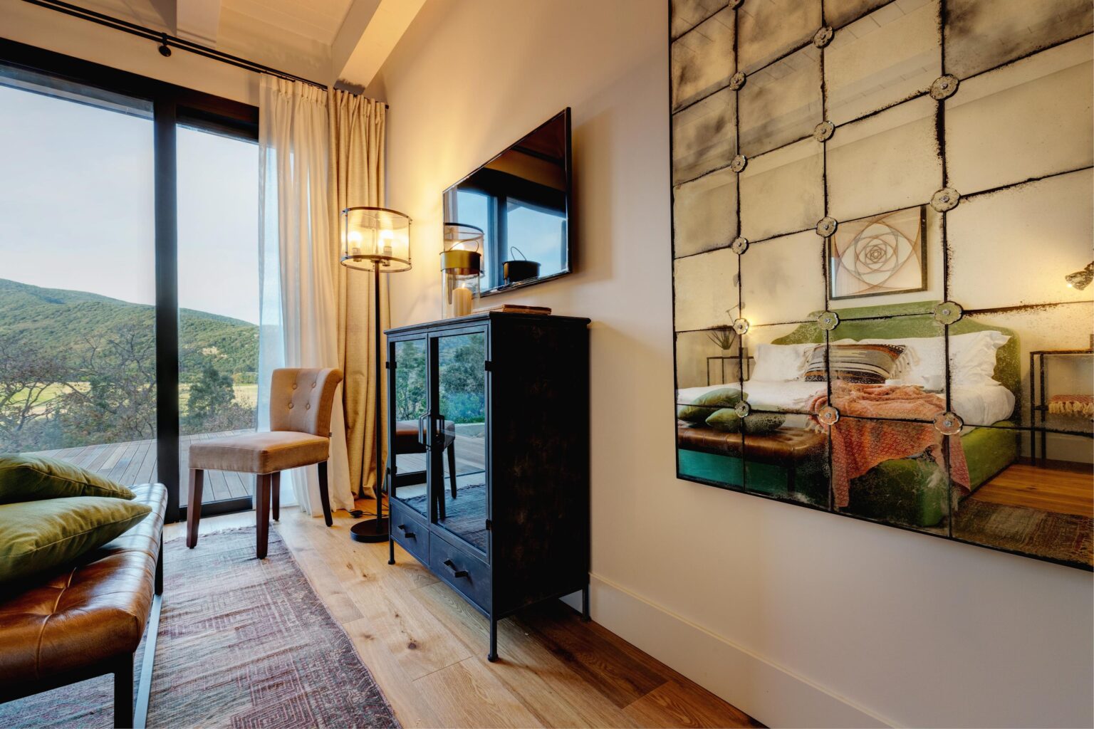 Cozy interior of a modern living room featuring a leather sofa, a stylish chair, and a black cabinet with glass doors. A large window offers a view of green mountains, while a decorative mirror reflects a vibrant bedroom with a green bed and colorful textiles. Soft lighting from a floor lamp enhances the warm ambiance. | Only on SopranoVillas