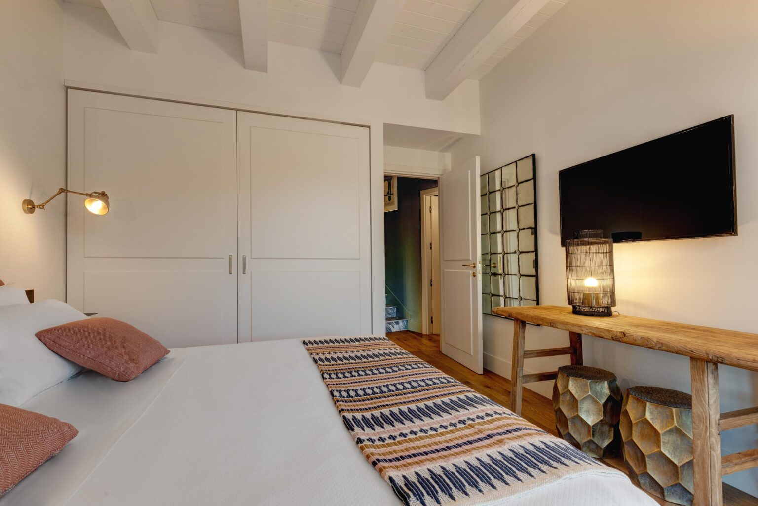 Cozy and modern bedroom featuring a queen-sized bed with decorative pillows, a stylish woven throw, a wooden console table, and a flat-screen TV. The room includes a large closet, a decorative mirror, and warm lighting, creating a welcoming atmosphere. Perfect for relaxation and comfort. | Only on SopranoVillas