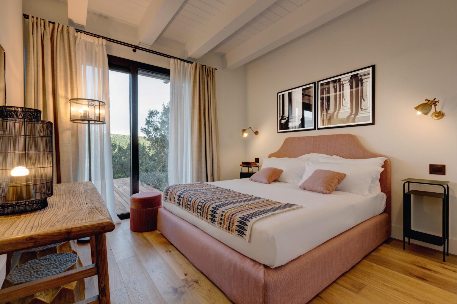 Cozy modern bedroom featuring a pink upholstered bed with decorative pillows, wooden flooring, and large windows for natural light. The room includes a stylish bedside table, a woven throw blanket, and framed artwork on the wall, creating a warm and inviting atmosphere. | Only on SopranoVillas