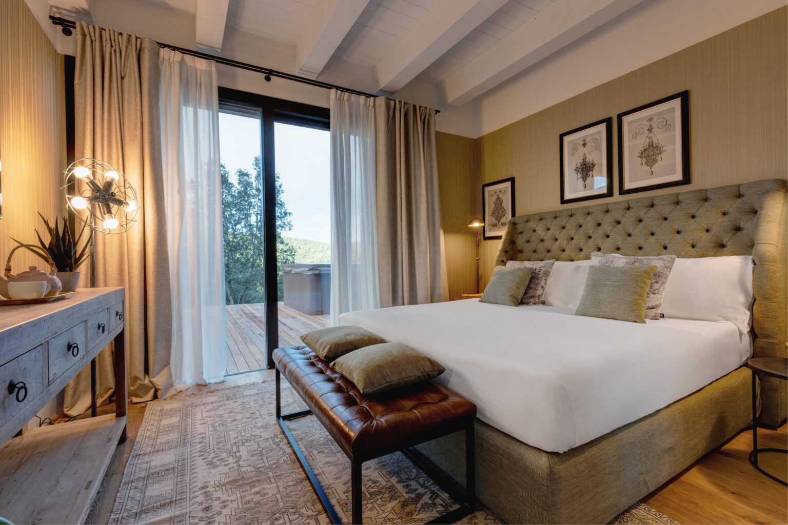 Modern bedroom interior featuring a plush bed with decorative pillows, a stylish leather bench, and a wooden dresser. Large glass doors lead to a balcony with scenic views. Soft lighting and elegant decor enhance the inviting atmosphere. | Only on SopranoVillas