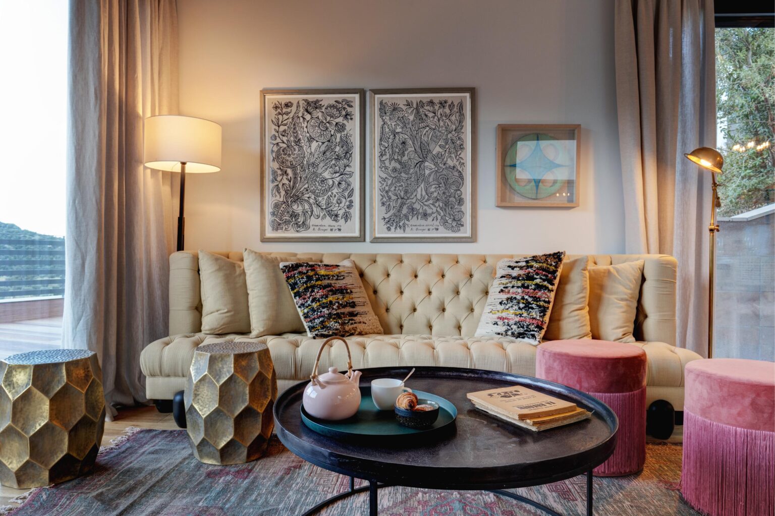 Modern living room featuring a tufted beige sofa adorned with colorful throw pillows, two stylish hexagonal gold side tables, and a round black coffee table holding a pink teapot, cup, pastry, and books. The walls are decorated with framed botanical artwork and a geometric print, complemented by natural light filtering through sheer curtains. | Only on SopranoVillas