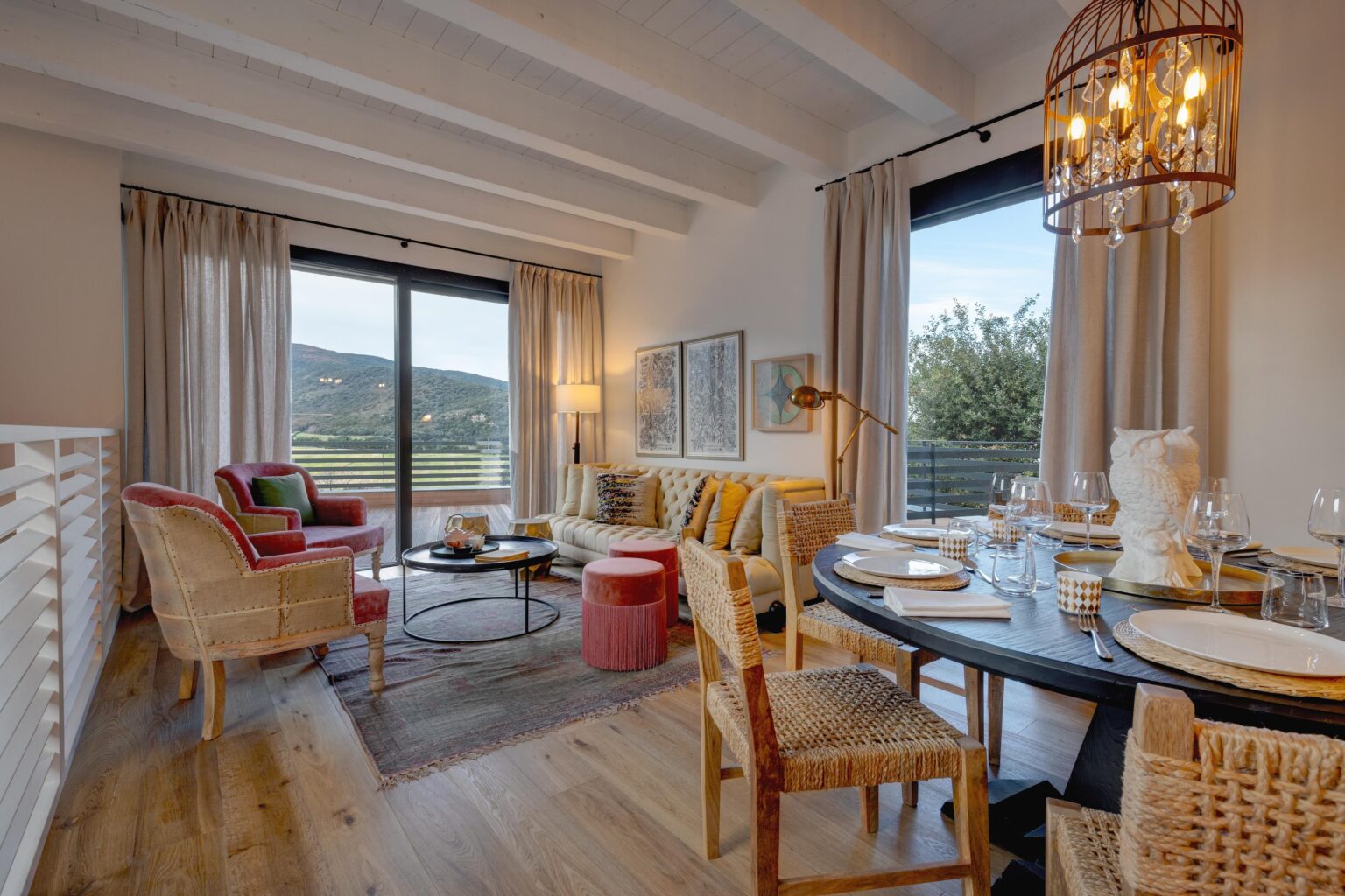 Cozy modern living room featuring a stylish sofa, colorful accent chairs, and a dining area with a round table set for dinner. Large windows provide scenic views of the outdoors, complemented by elegant decor and warm lighting. Ideal for showcasing contemporary interior design. | Only on SopranoVillas