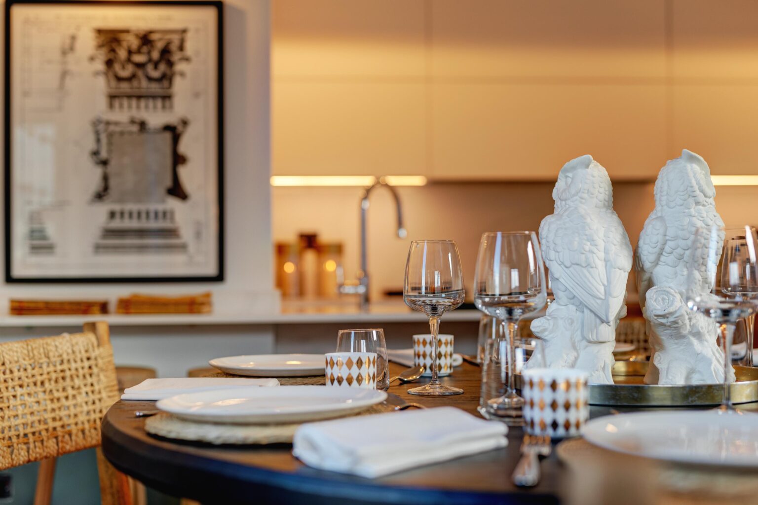 Elegant dining table set with white plates, decorative owl figurines, and various glassware, featuring a modern kitchen backdrop and artistic wall decor. | Only on SopranoVillas