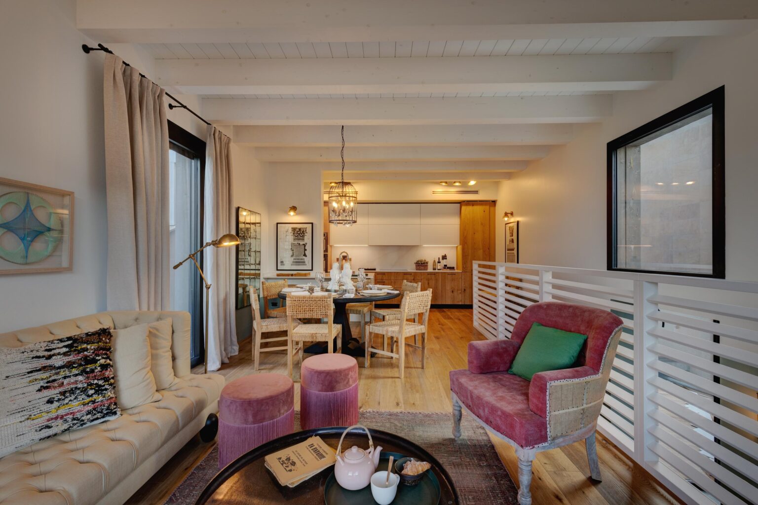 Modern living room and dining area featuring a beige tufted sofa, pink ottomans, and a wooden dining table set for four. The space has white beams, large windows with curtains, and stylish decor elements including artwork and a chandelier. | Only on SopranoVillas