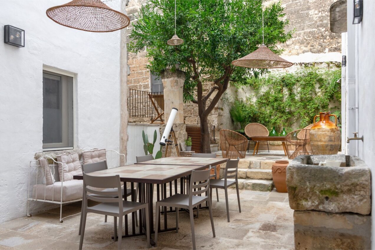 Charming outdoor patio featuring a dining table with modern chairs, a cozy seating area, and lush greenery. The space is enhanced by decorative hanging lights and a rustic stone wall, creating a serene atmosphere for relaxation and dining. | Only on SopranoVillas