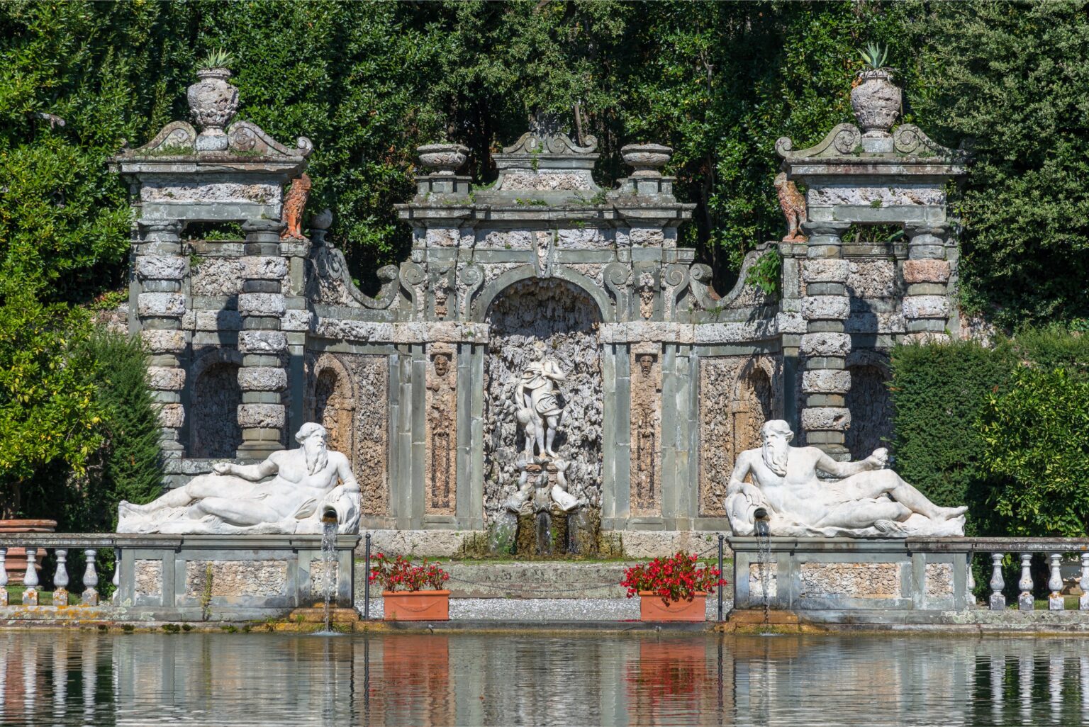 Baroque-style fountain with intricate stonework and sculptures, featuring reclining figures and a central water feature, surrounded by lush greenery and reflecting in the water. | Only on SopranoVillas