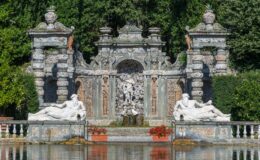 Baroque-style fountain with intricate stonework and sculptures, featuring reclining figures and a central water feature, surrounded by lush greenery and reflecting in the water.