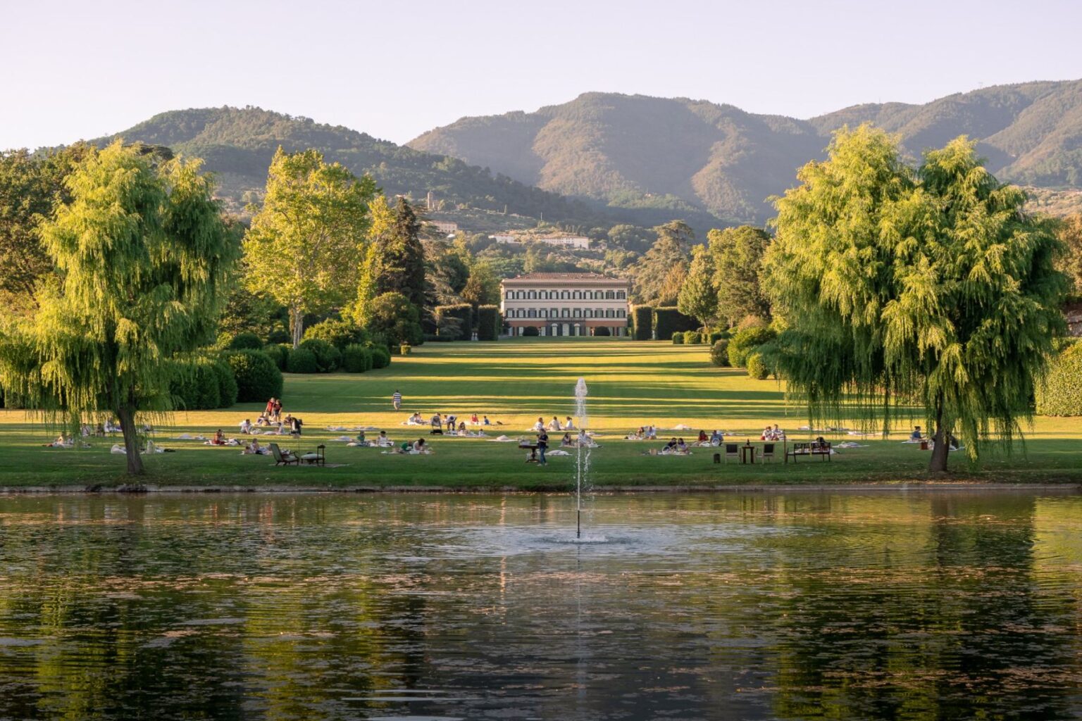 Scenic park view featuring lush green lawns, a tranquil pond with a fountain, and people enjoying leisure activities. The background showcases a historic building framed by mountains, creating a serene outdoor atmosphere ideal for relaxation and picnics. | Only on SopranoVillas