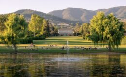 Scenic park view featuring lush green lawns, a tranquil pond with a fountain, and people enjoying leisure activities. The background showcases a historic building framed by mountains, creating a serene outdoor atmosphere ideal for relaxation and picnics.