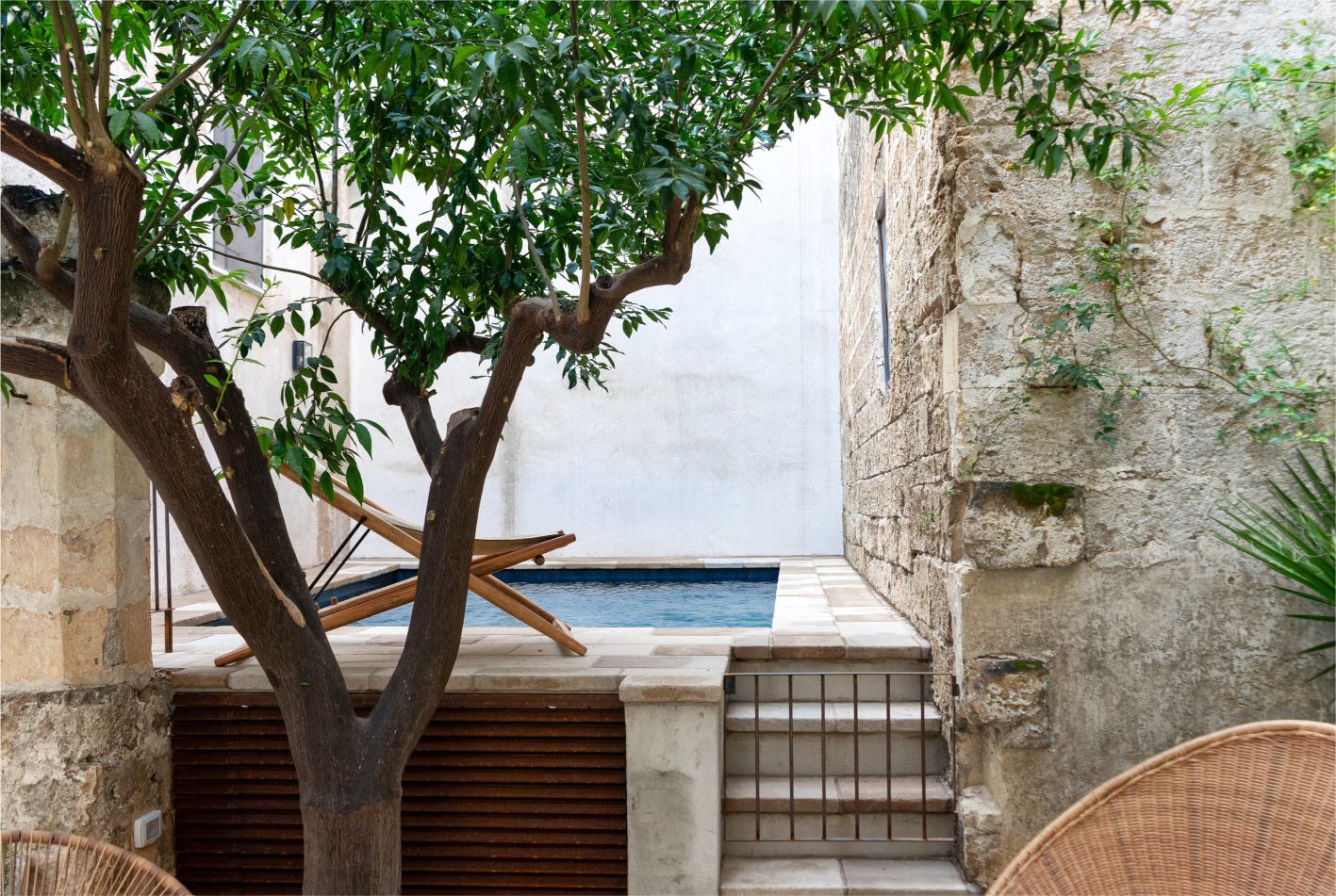 A serene courtyard featuring a lush tree, a wooden lounge chair, and a small swimming pool surrounded by stone walls, creating a tranquil outdoor space ideal for relaxation. | Only on SopranoVillas