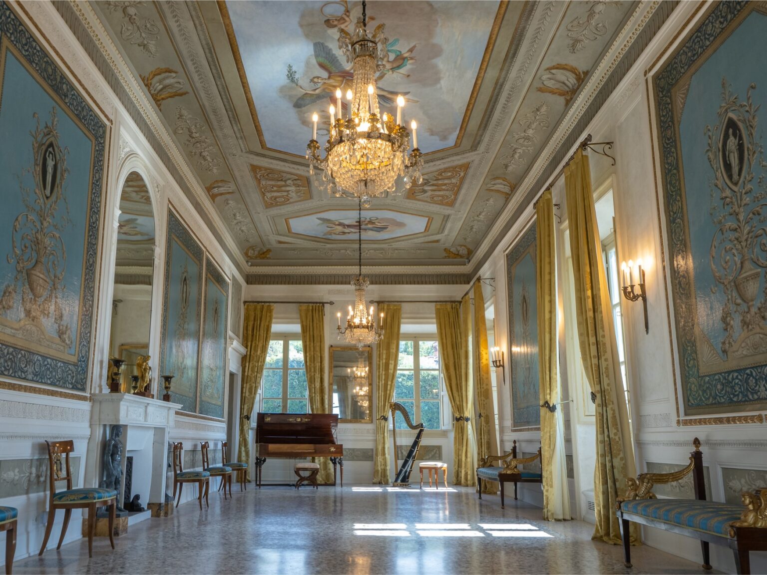 Elegant interior of a grand hall featuring ornate blue and gold wall decorations, a chandelier, large windows with yellow curtains, and a piano. The space showcases intricate ceiling artwork and classic furnishings, creating a luxurious ambiance. | Only on SopranoVillas