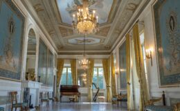 Elegant interior of a grand hall featuring ornate blue and gold wall decorations, a chandelier, large windows with yellow curtains, and a piano. The space showcases intricate ceiling artwork and classic furnishings, creating a luxurious ambiance.