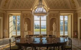 Elegant dining room featuring a round wooden table surrounded by chairs, adorned with decorative elements. The room is illuminated by a chandelier and showcases classical sculptures positioned near large windows, offering a view of the gardens outside. The ceiling features intricate designs, enhancing the room's historical charm.