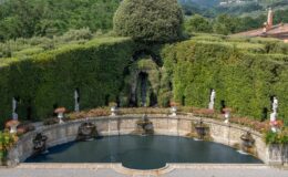 Lush garden with a circular pond surrounded by manicured hedges, featuring ornate fountains and classical statues, set against a backdrop of rolling hills and greenery.