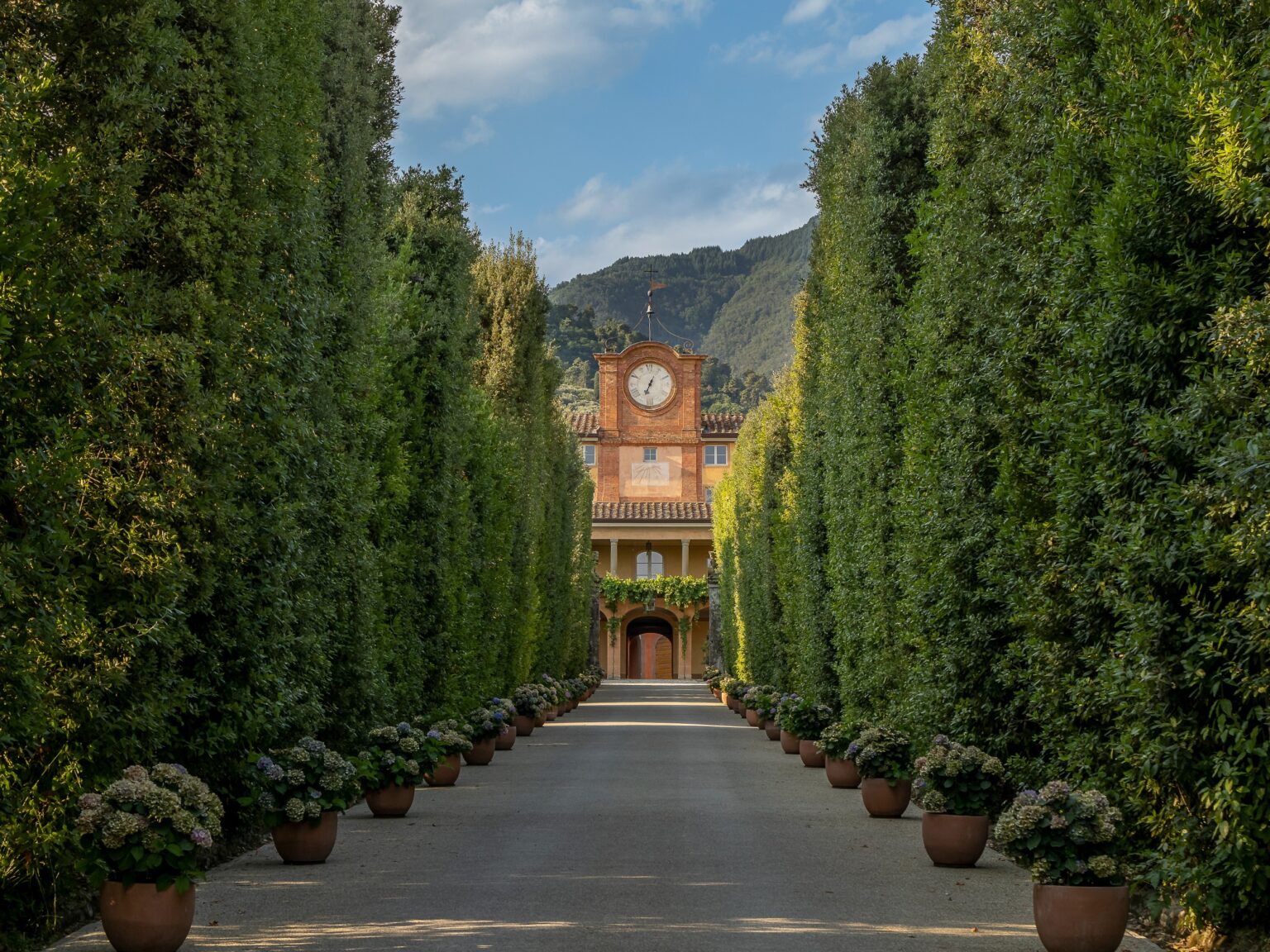 A picturesque garden pathway lined with tall hedges and potted flowers, leading to a historic building with a clock tower, set against a backdrop of mountains and blue skies. | Only on SopranoVillas