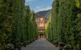 A picturesque garden pathway lined with tall hedges and potted flowers, leading to a historic building with a clock tower, set against a backdrop of mountains and blue skies.