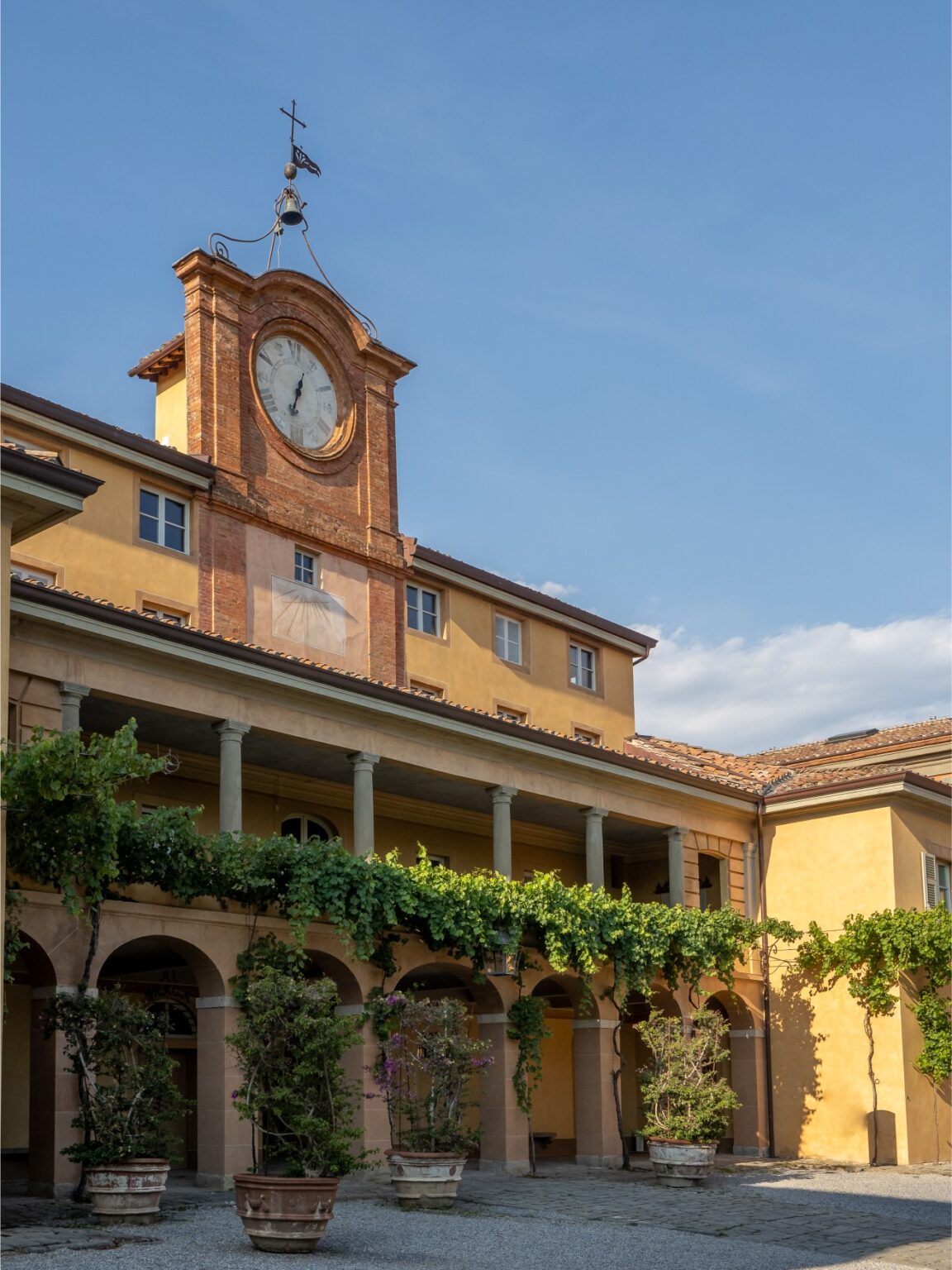 Historic building with a prominent clock tower, featuring a warm yellow facade and arched columns adorned with greenery and potted plants, set against a clear blue sky. | Only on SopranoVillas