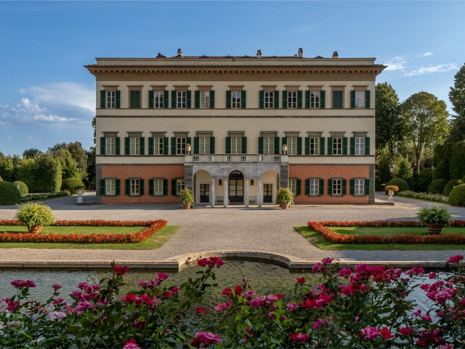 Elegant villa with green shutters surrounded by manicured gardens and colorful flower beds, reflecting classic architectural design under a clear blue sky. | Only on SopranoVillas