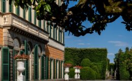 Elegant historic villa exterior with green shutters, detailed balcony, and manicured garden featuring ornamental planters and hedges under a clear blue sky.