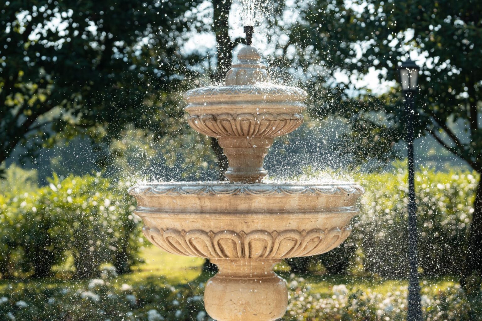 Elegant three-tiered stone fountain with water cascading and sparkling in sunlight, surrounded by lush greenery and soft bokeh effect. | Only on SopranoVillas