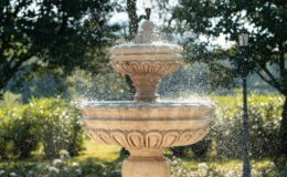 Elegant three-tiered stone fountain with water cascading and sparkling in sunlight, surrounded by lush greenery and soft bokeh effect.