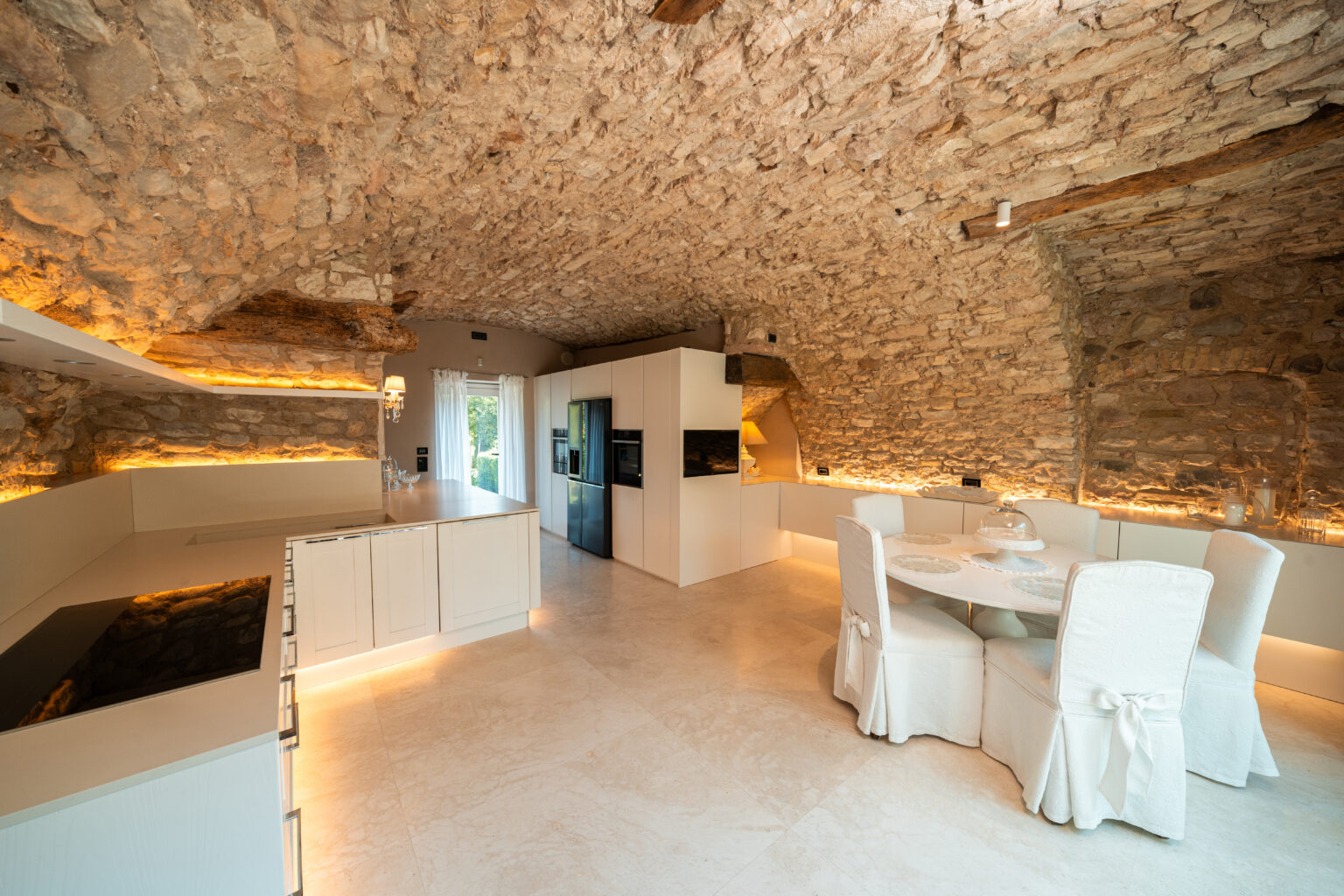 Modern kitchen interior with stone walls and a curved ceiling, featuring sleek white cabinetry, a minimalist dining table with upholstered chairs, and warm ambient lighting. Natural light streams through large windows, enhancing the inviting atmosphere. | Only on SopranoVillas