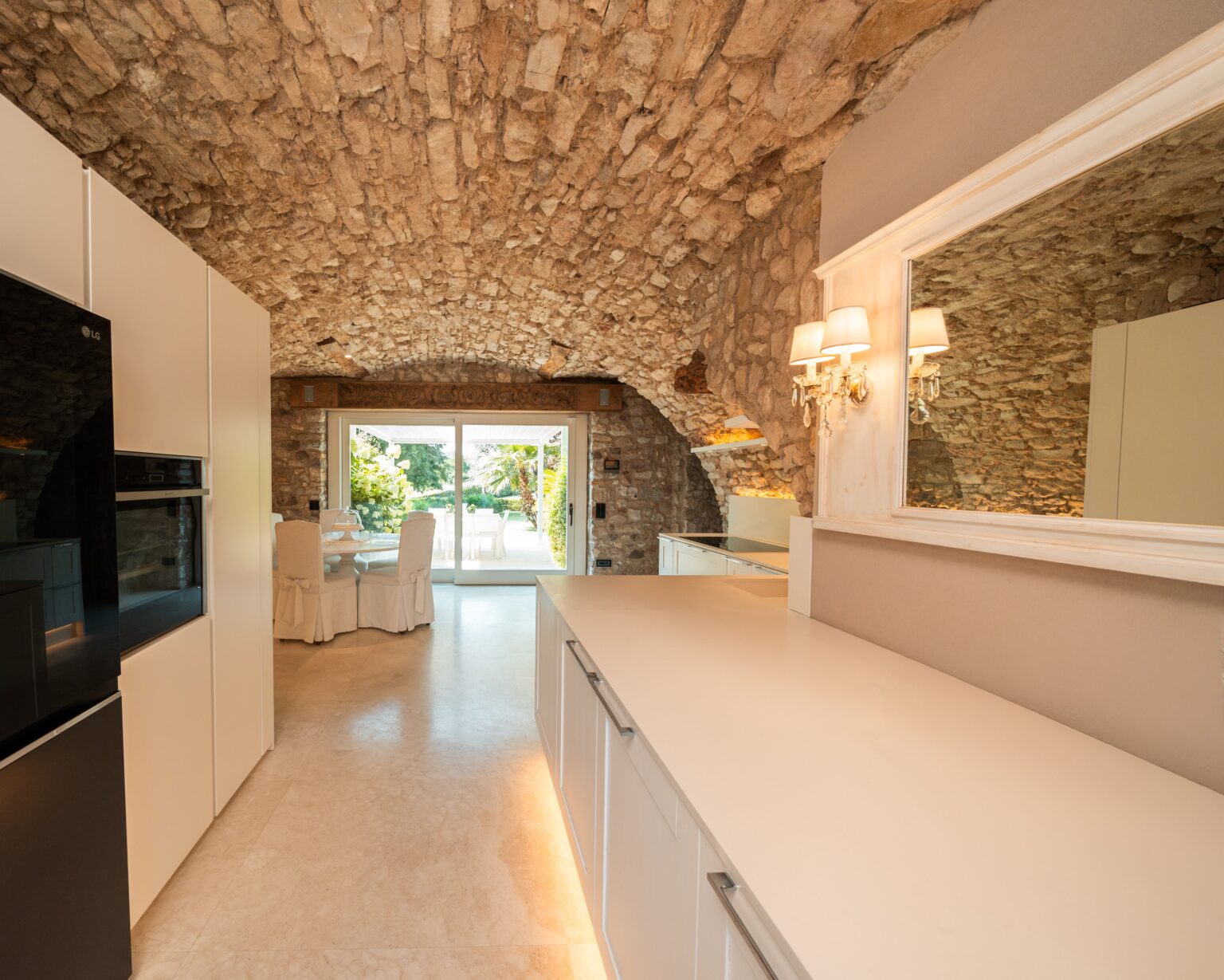 Modern kitchen with a stone arched ceiling, featuring white cabinetry, a sleek countertop, and a view of an outdoor dining area through large sliding glass doors. Natural light illuminates the space, enhancing the warm and inviting atmosphere. | Only on SopranoVillas