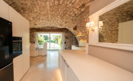 Modern kitchen with a stone arched ceiling, featuring white cabinetry, a sleek countertop, and a view of an outdoor dining area through large sliding glass doors. Natural light illuminates the space, enhancing the warm and inviting atmosphere.
