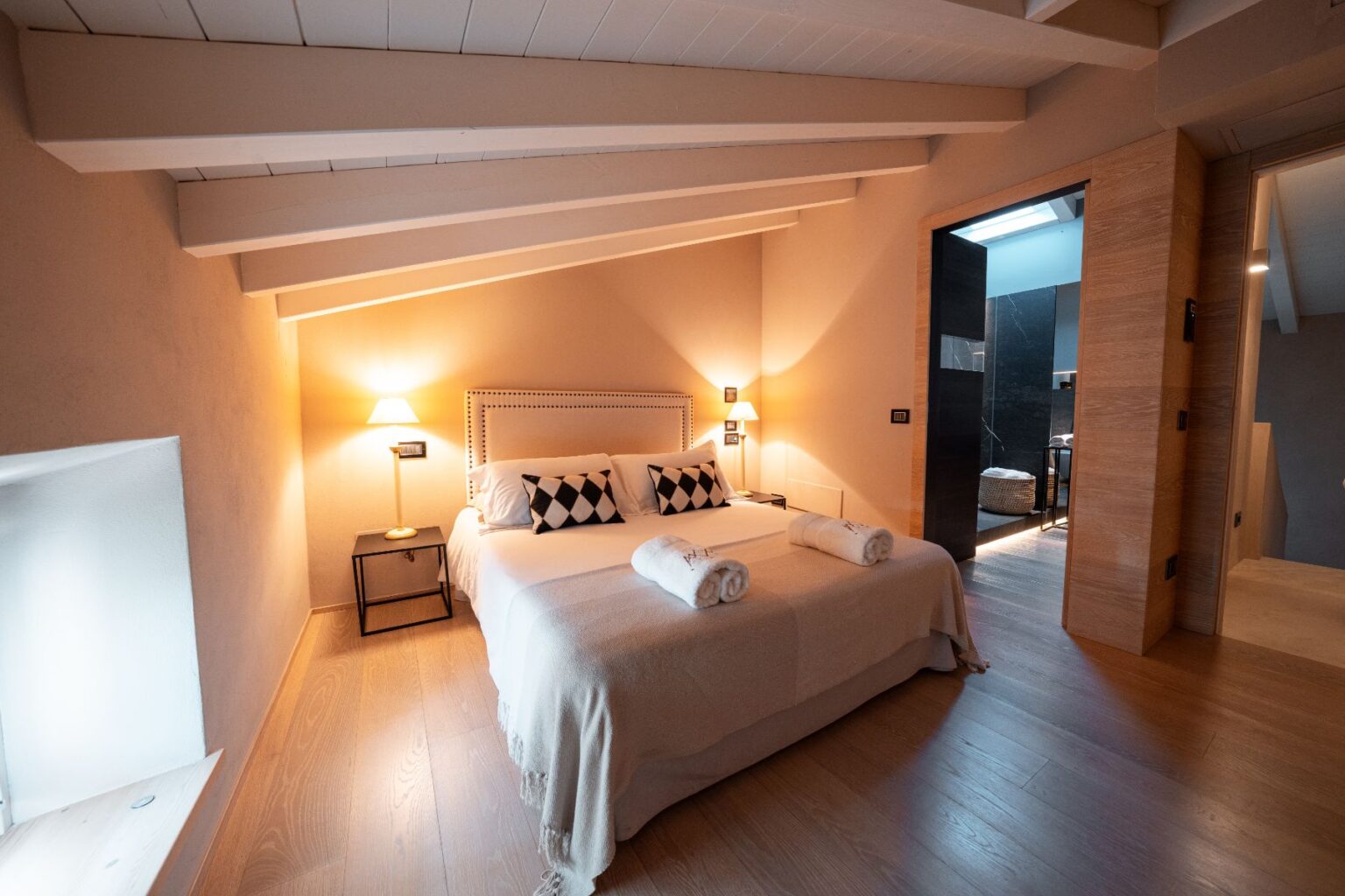 Cozy bedroom featuring a queen-sized bed with decorative pillows, soft towels, and warm lighting, complemented by wooden flooring and a stylish en-suite bathroom entrance. Ideal for a relaxing getaway. | Only on SopranoVillas