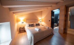 Cozy bedroom featuring a queen-sized bed with decorative pillows, soft towels, and warm lighting, complemented by wooden flooring and a stylish en-suite bathroom entrance. Ideal for a relaxing getaway.