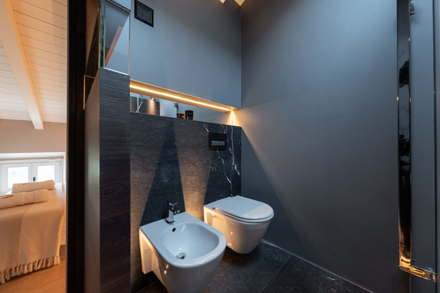 Modern bathroom featuring a sleek toilet and bidet against a dark stone wall, illuminated by warm LED lighting. The minimalist design is complemented by a stylish gray color palette and wooden accents, creating a sophisticated atmosphere. | Only on SopranoVillas