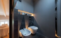Modern bathroom featuring a sleek toilet and bidet against a dark stone wall, illuminated by warm LED lighting. The minimalist design is complemented by a stylish gray color palette and wooden accents, creating a sophisticated atmosphere.