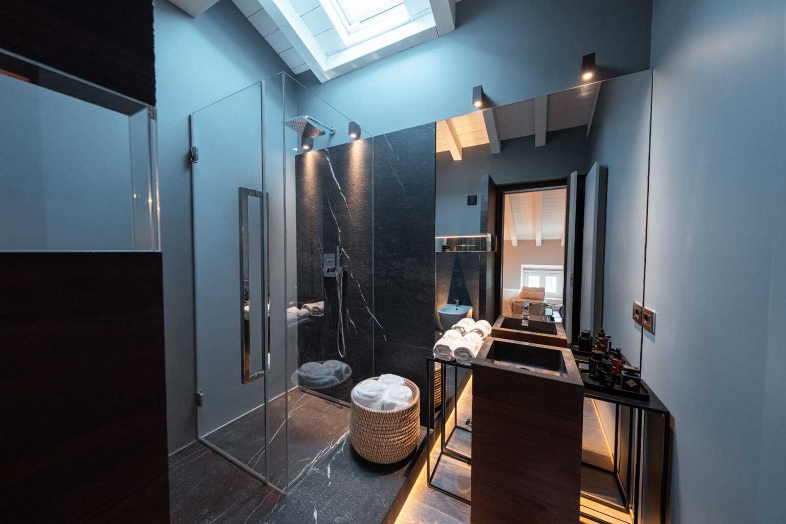 Modern bathroom featuring a glass shower enclosure, sleek black marble walls, and stylish wooden vanities. The space is illuminated by natural light from a skylight, showcasing neatly stacked towels and elegant bathroom accessories. | Only on SopranoVillas
