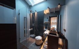 Modern bathroom featuring a glass shower enclosure, sleek black marble walls, and stylish wooden vanities. The space is illuminated by natural light from a skylight, showcasing neatly stacked towels and elegant bathroom accessories.