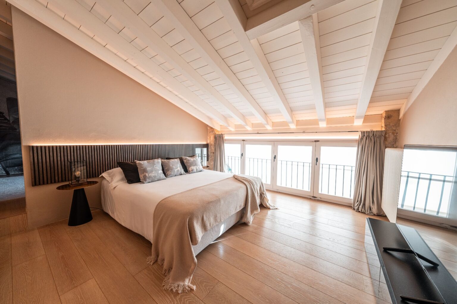 Modern bedroom with wooden flooring, a cozy double bed adorned with decorative pillows, and a soft throw blanket. The room features sloped white beams and large windows with curtains, allowing ample natural light. A stylish side table and a sleek TV unit complete the contemporary decor. Ideal for relaxation and comfort. | Only on SopranoVillas