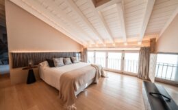 Modern bedroom with wooden flooring, a cozy double bed adorned with decorative pillows, and a soft throw blanket. The room features sloped white beams and large windows with curtains, allowing ample natural light. A stylish side table and a sleek TV unit complete the contemporary decor. Ideal for relaxation and comfort.