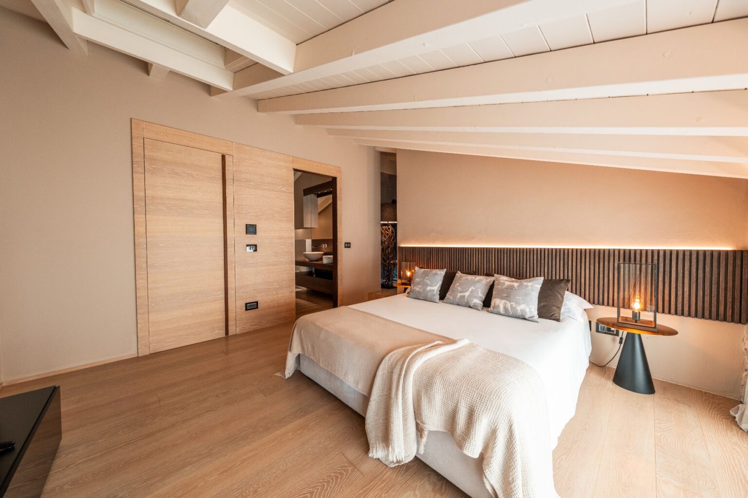 Modern bedroom interior featuring a cozy bed with decorative pillows, a stylish bedside table with a lamp, and warm lighting. The design includes wooden accents, neutral color tones, and a spacious layout, creating a serene and inviting atmosphere. Ideal for relaxation and comfort. | Only on SopranoVillas