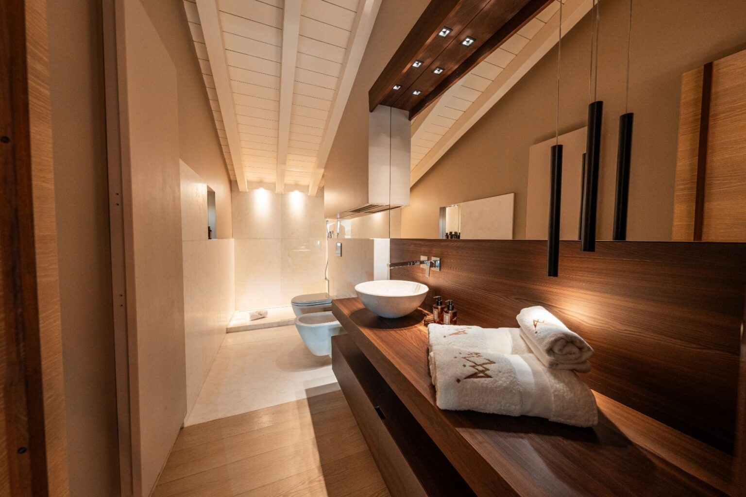 Modern bathroom interior featuring a stylish wooden countertop with a round basin, neatly arranged towels, and elegant lighting. The design includes a spacious layout with a combination of wooden and marble finishes, creating a serene and contemporary atmosphere. | Only on SopranoVillas