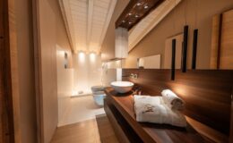 Modern bathroom interior featuring a stylish wooden countertop with a round basin, neatly arranged towels, and elegant lighting. The design includes a spacious layout with a combination of wooden and marble finishes, creating a serene and contemporary atmosphere.