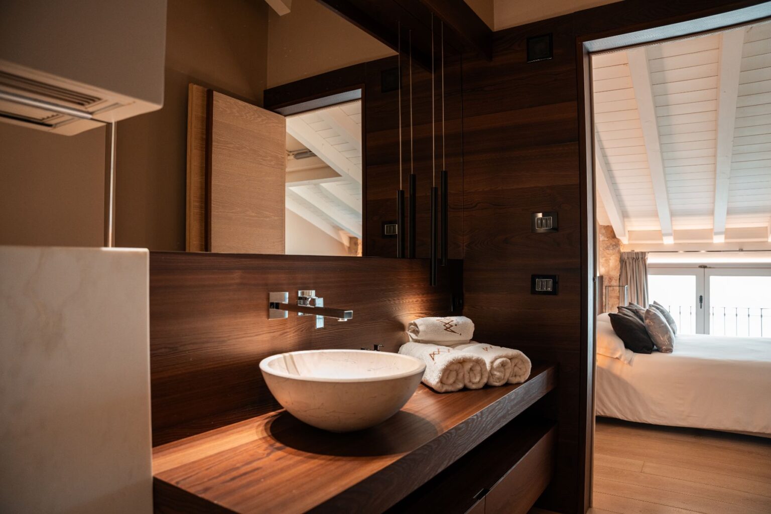 Modern bathroom design featuring a stylish wooden vanity with a round marble sink, neatly stacked white towels, and elegant lighting fixtures. A glimpse of a cozy bedroom with a large window and soft bedding is visible in the background, highlighting a harmonious blend of functionality and aesthetics. | Only on SopranoVillas