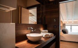 Modern bathroom design featuring a stylish wooden vanity with a round marble sink, neatly stacked white towels, and elegant lighting fixtures. A glimpse of a cozy bedroom with a large window and soft bedding is visible in the background, highlighting a harmonious blend of functionality and aesthetics.