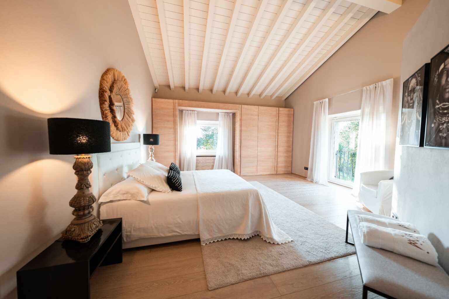Spacious modern bedroom featuring a king-size bed with elegant bedding, decorative pillows, stylish lamps, and large windows that provide natural light. The room has a light color palette, wooden flooring, and a cozy seating area, creating a serene and inviting atmosphere. | Only on SopranoVillas