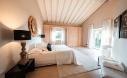 Spacious modern bedroom featuring a king-size bed with elegant bedding, decorative pillows, stylish lamps, and large windows that provide natural light. The room has a light color palette, wooden flooring, and a cozy seating area, creating a serene and inviting atmosphere.