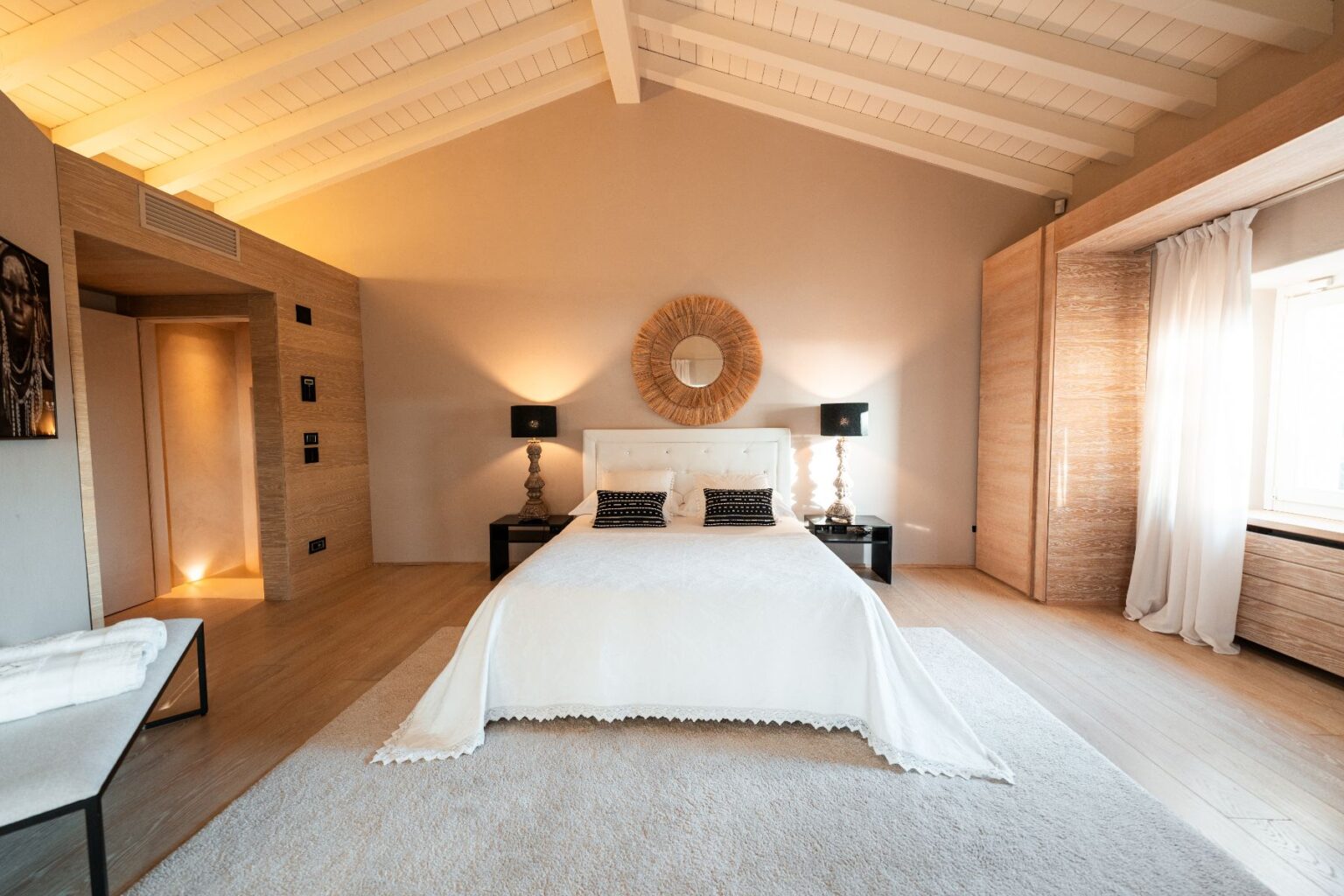 Modern bedroom featuring a king-size bed with a white coverlet, decorative black pillows, and stylish bedside lamps. The room has high ceilings with wooden beams, a round mirror above the bed, and large windows with sheer curtains. Warm lighting and minimalist decor create a cozy atmosphere. | Only on SopranoVillas