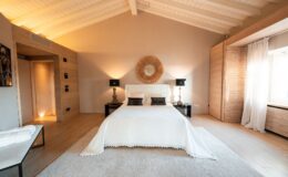Modern bedroom featuring a king-size bed with a white coverlet, decorative black pillows, and stylish bedside lamps. The room has high ceilings with wooden beams, a round mirror above the bed, and large windows with sheer curtains. Warm lighting and minimalist decor create a cozy atmosphere.