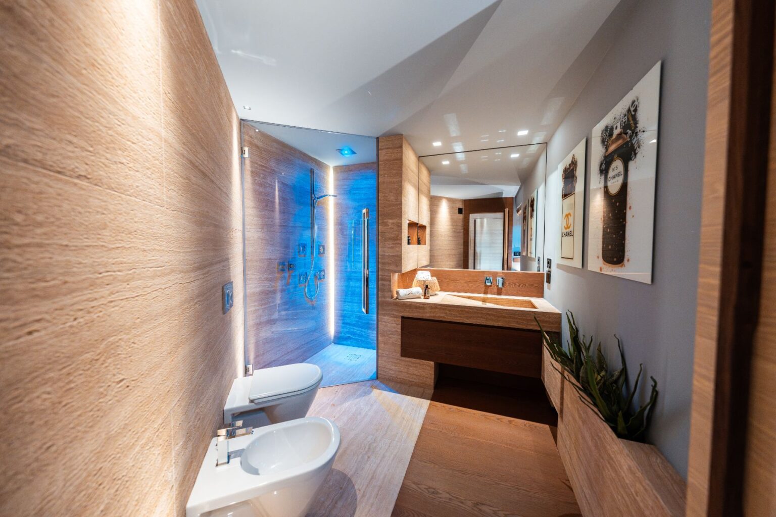 Modern bathroom interior featuring wooden accents, a spacious shower with blue lighting, dual toilets, and a sleek vanity with a minimalist design. The walls are adorned with decorative art, enhancing the contemporary aesthetic. | Only on SopranoVillas