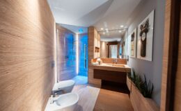 Modern bathroom interior featuring wooden accents, a spacious shower with blue lighting, dual toilets, and a sleek vanity with a minimalist design. The walls are adorned with decorative art, enhancing the contemporary aesthetic.