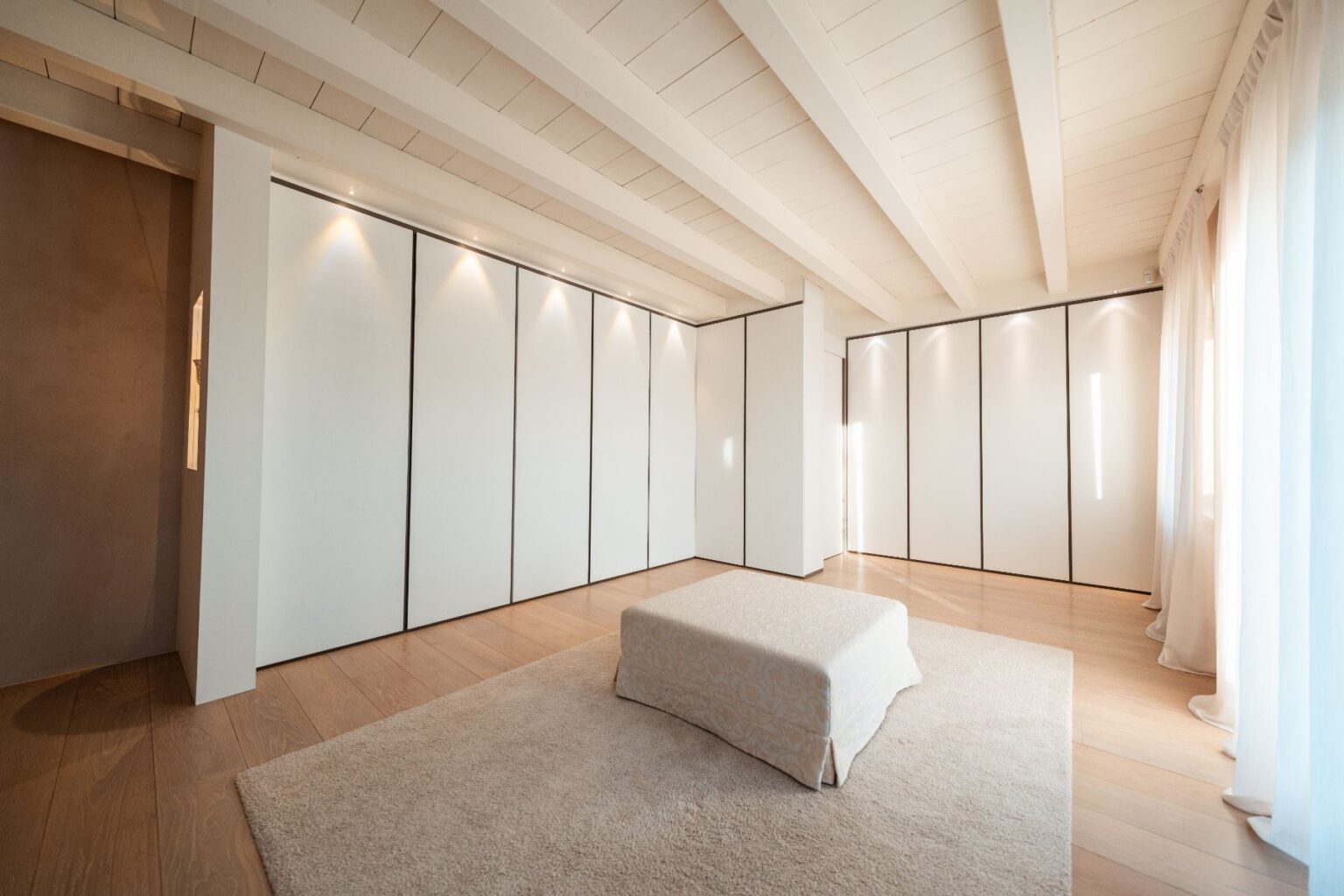 Modern minimalist bedroom interior featuring white panel walls, wooden flooring, and soft natural lighting. A plush area rug complements a low, upholstered bed in the center, creating a serene and spacious atmosphere. | Only on SopranoVillas