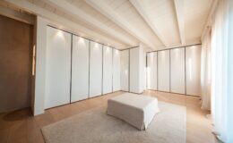 Modern minimalist bedroom interior featuring white panel walls, wooden flooring, and soft natural lighting. A plush area rug complements a low, upholstered bed in the center, creating a serene and spacious atmosphere.