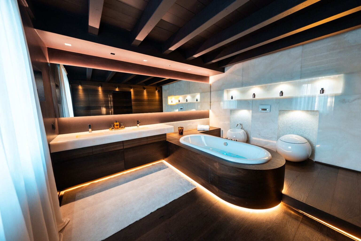 Modern luxury bathroom featuring a freestanding tub, dual sinks, and ambient LED lighting. The design incorporates natural wood and stone elements, with large windows allowing natural light to enhance the serene atmosphere. | Only on SopranoVillas
